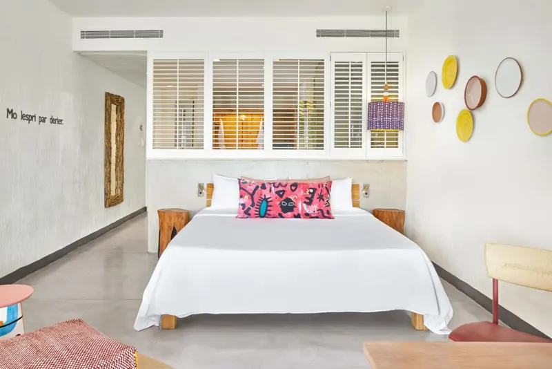 Une chambre lumineuse avec un grand lit recouvert d'une courtepointe blanche et de coussins colorés.