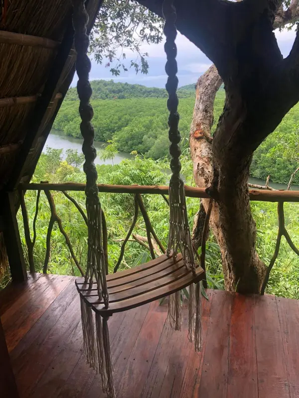Une balancelle en macramé se trouve sur une terrasse en bois avec vue sur une végétation luxuriante.