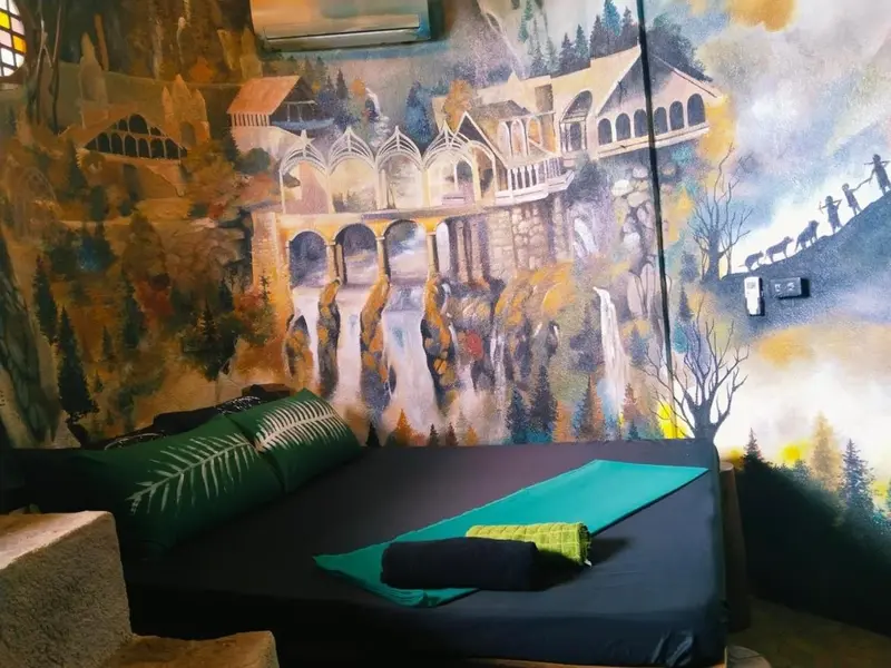 Une chambre avec un lit recouvert d'une couverture sombre et de coussins verts, face à un mur peint d'un paysage urbain.