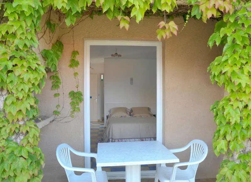 Une terrasse ombragée donne sur une chambre lumineuse avec un lit fait.