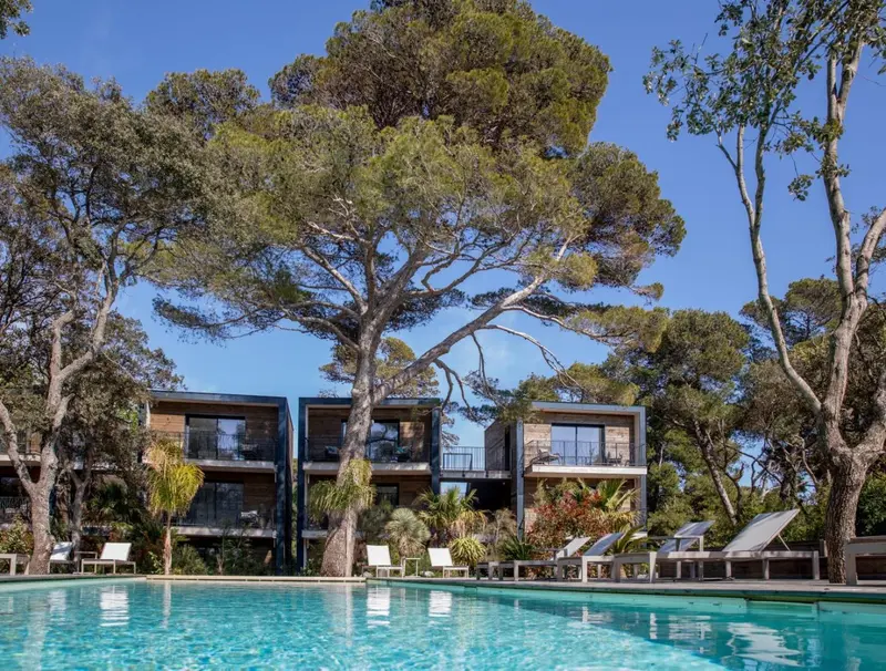Une piscine turquoise bordée de transats et de végétation luxuriante devant des bâtiments modernes.