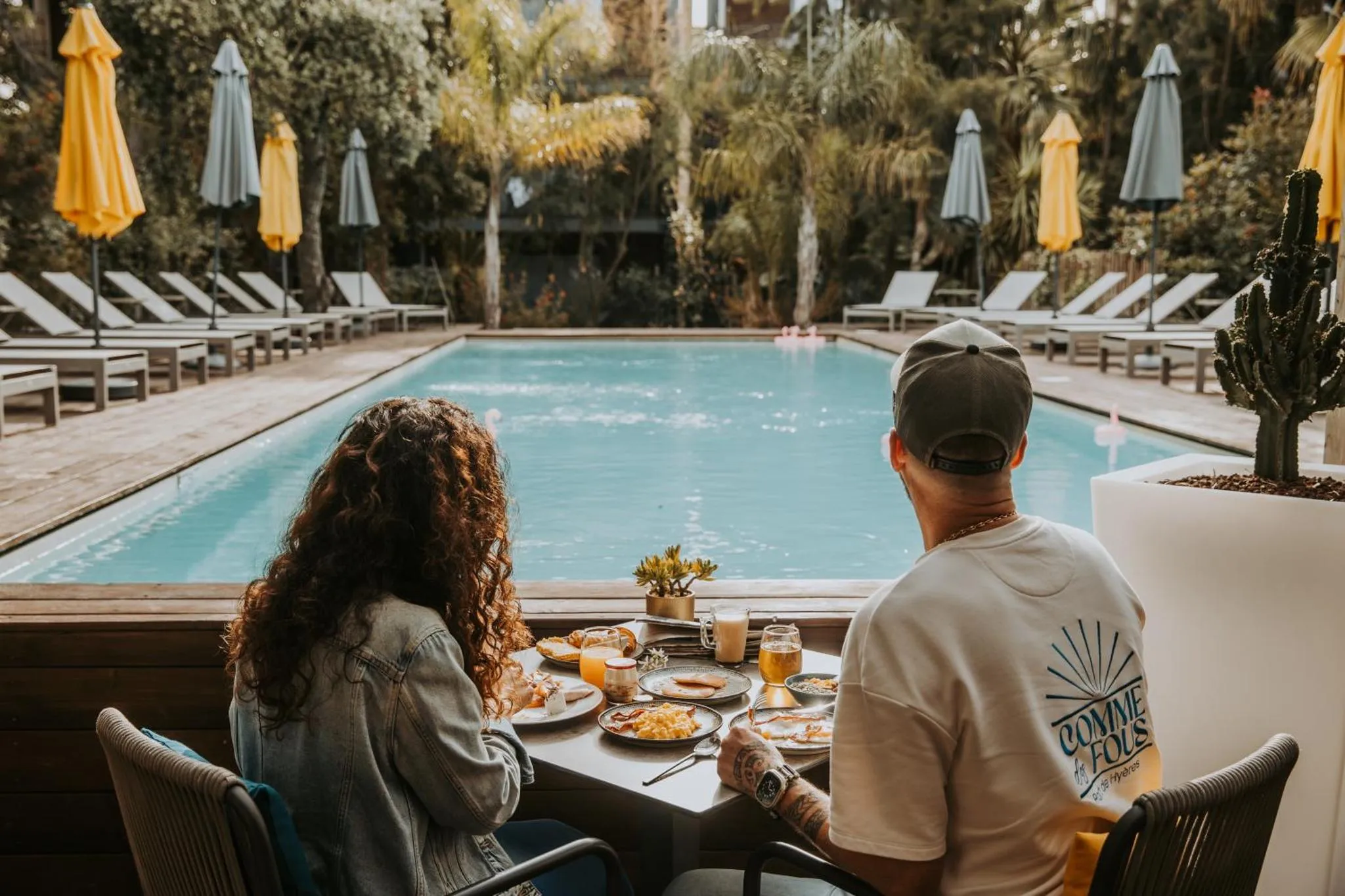 Deux personnes sont assises à une table en terrasse, face à une piscine.