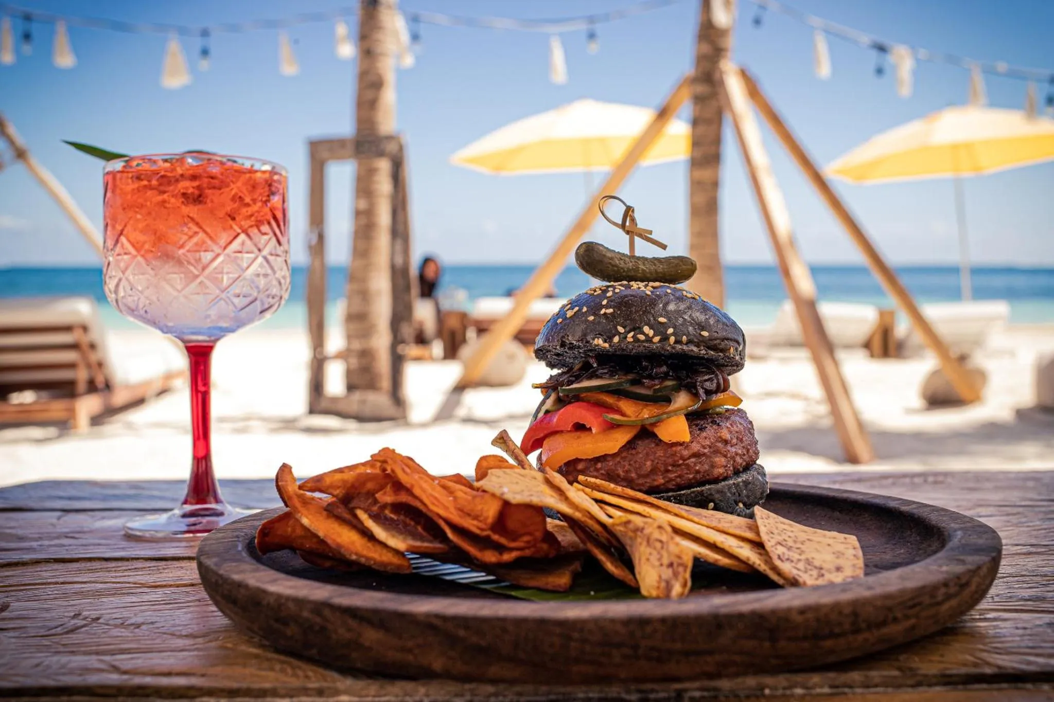 Un cocktail et un hamburger colorés sont servis sur une table en bois face à la mer.