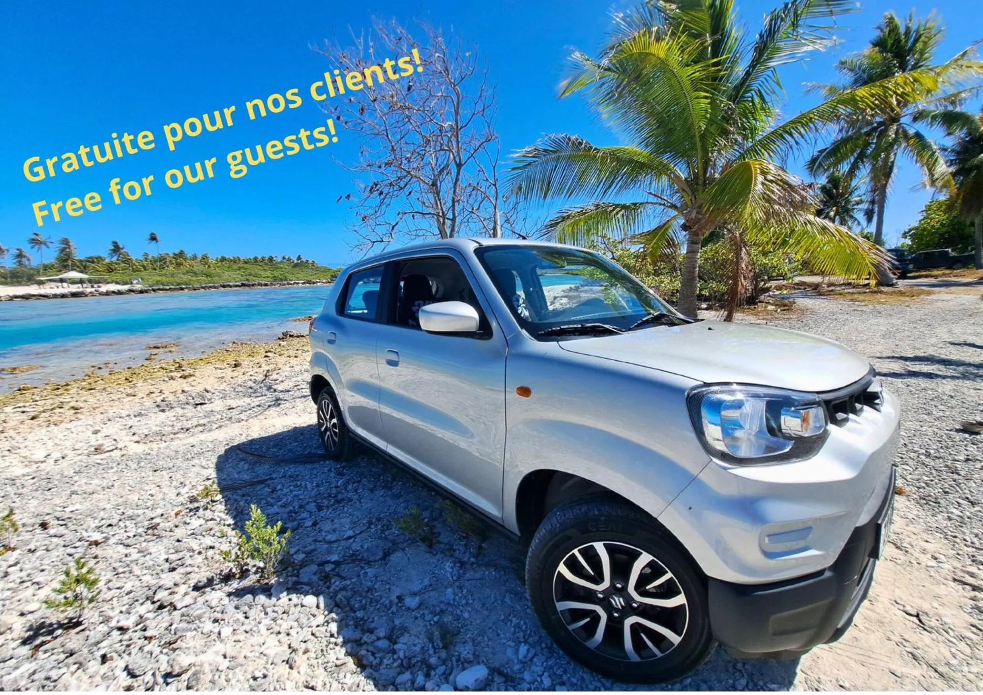 Une voiture argentée est garée sur un chemin de sable blanc bordé de végétation tropicale.