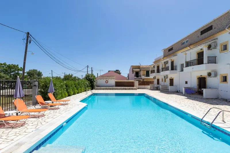 Une piscine rectangulaire bordée de chaises longues se trouve devant un bâtiment blanc à deux étages.