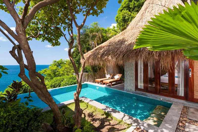 Une petite piscine turquoise est bordée de chaises longues devant un bungalow à toit de chaume.