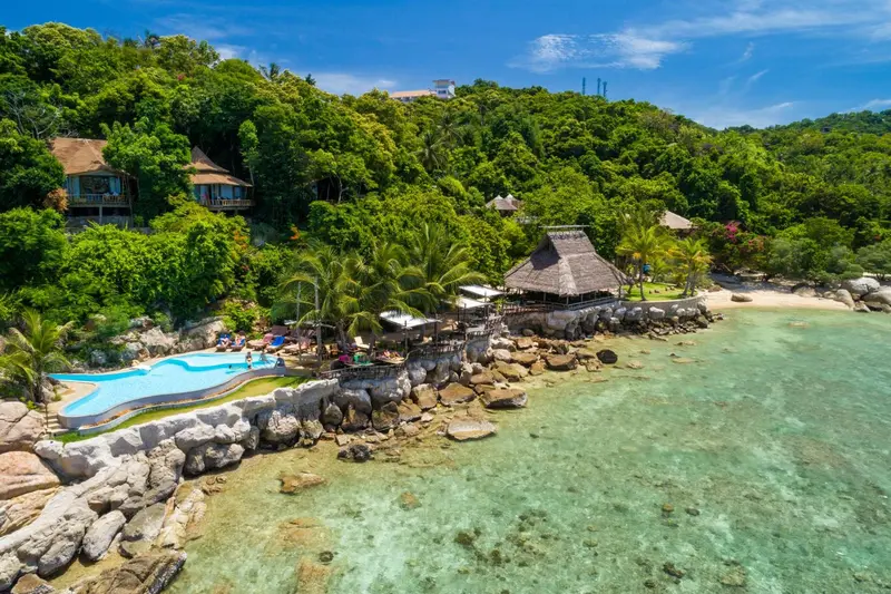 Vue aérienne d'une plage avec une piscine et des bungalows sur une colline verdoyante.