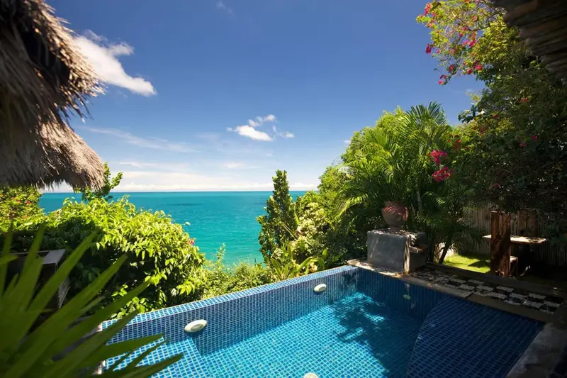 Petite piscine turquoise entourée de végétation luxuriante avec vue sur la mer.