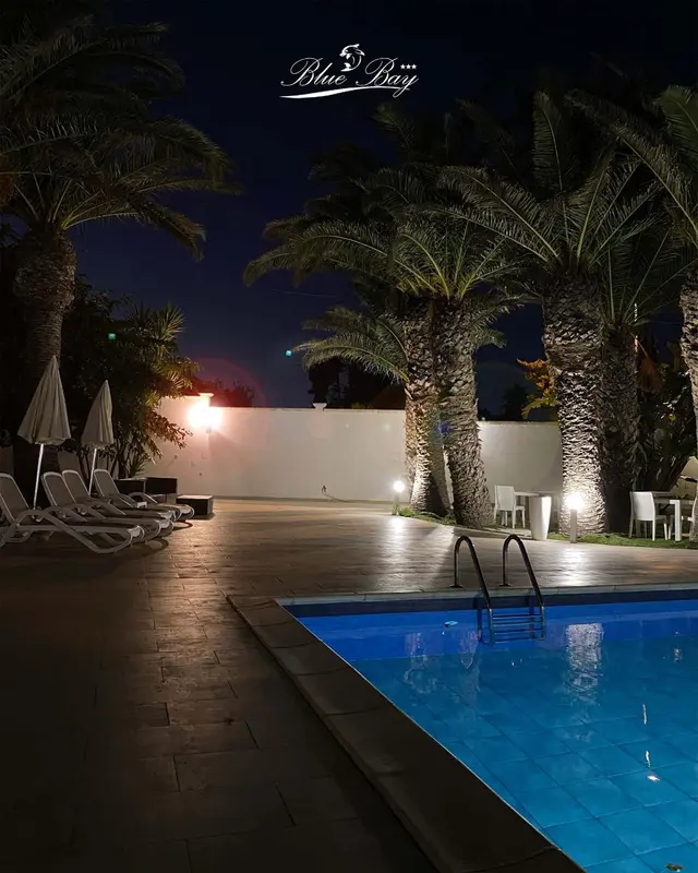 Terrasse lumineuse avec piscine et palmiers la nuit.