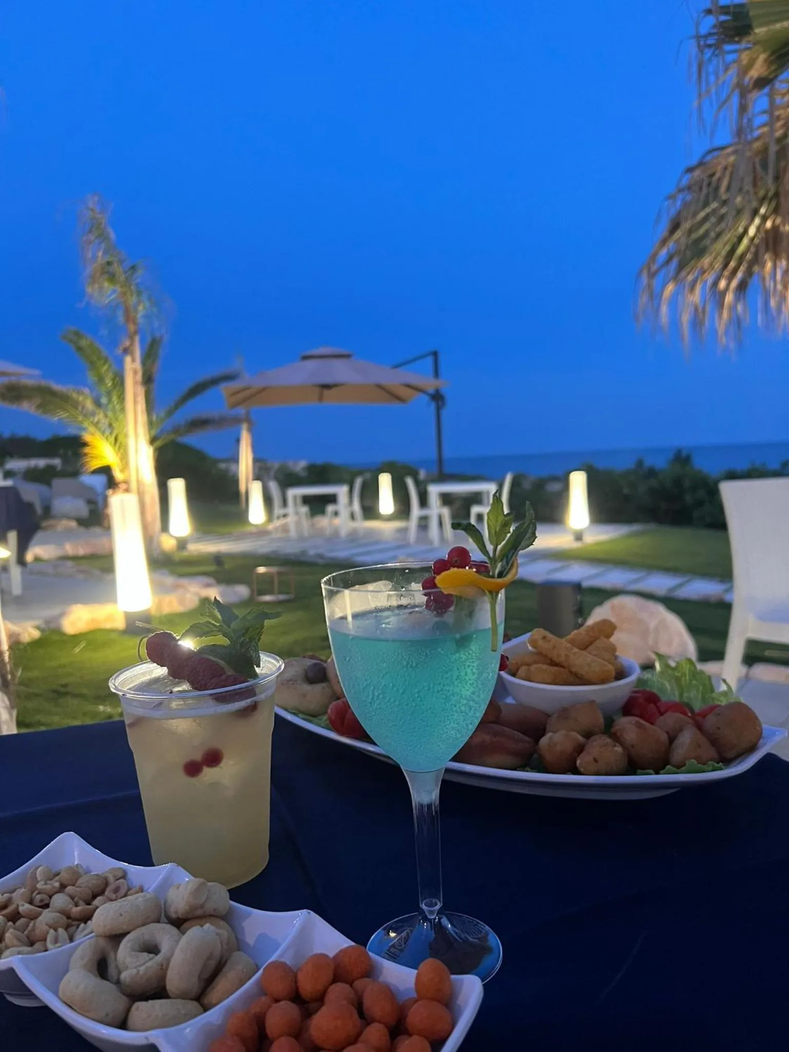 Une table dressée avec des boissons et des amuse-gueules en extérieur, face à la mer au crépuscule.