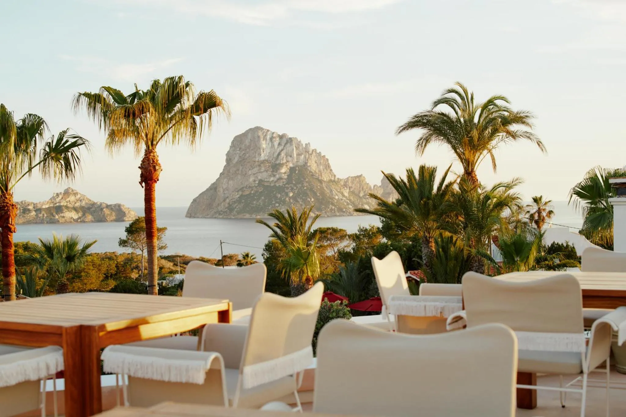 Tables et chaises claires sur une terrasse ensoleillée avec vue sur la mer et une formation rocheuse.