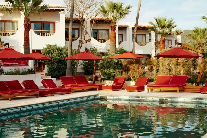 Transats rouges disposés autour d'une piscine turquoise, devant un bâtiment blanc orné de palmiers.