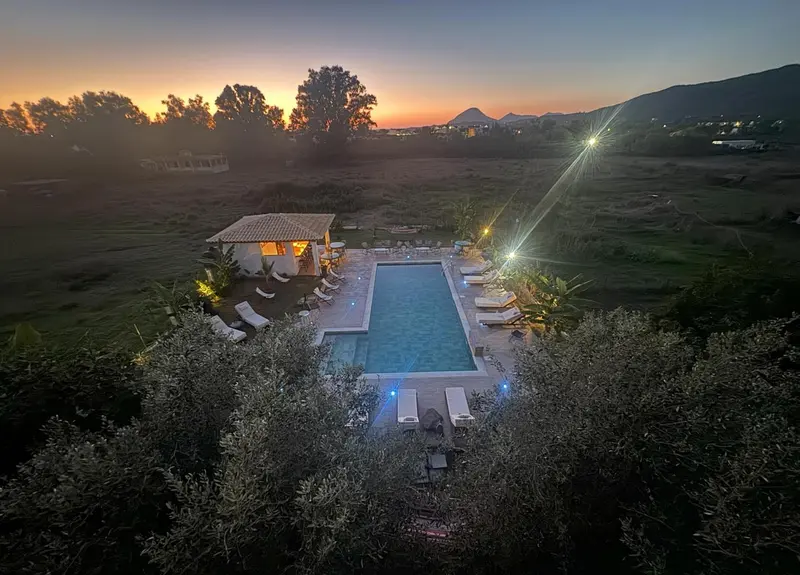 Vue aérienne d'une piscine illuminée au crépuscule, entourée de chaises longues et de végétation.