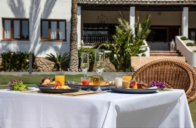 Une table dressée avec des petits déjeuners est mise en scène devant un bâtiment blanc et une végétation luxuriante.