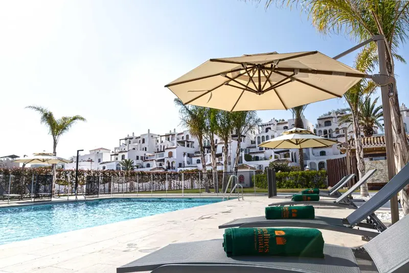 Transats verts et parasols au bord d'une piscine turquoise, avec des bâtiments blancs en arrière-plan.