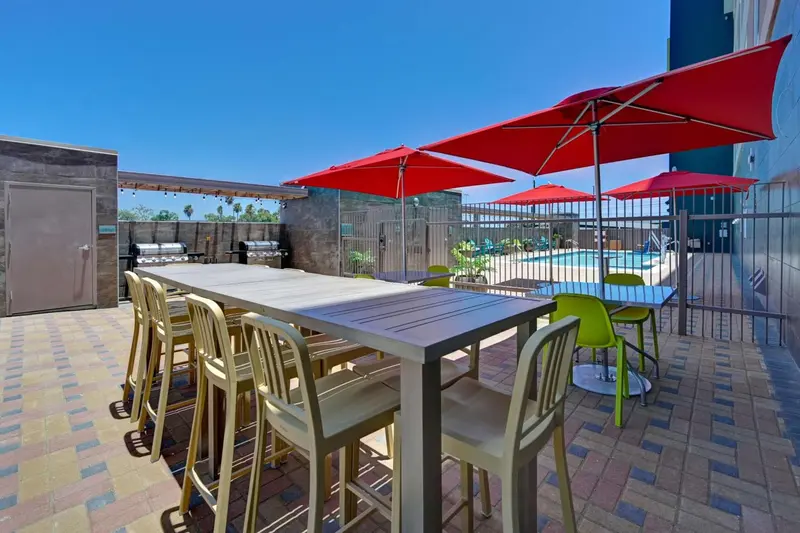 Une longue table et des chaises se trouvent sur une terrasse pavée, à l'ombre de parasols rouges.