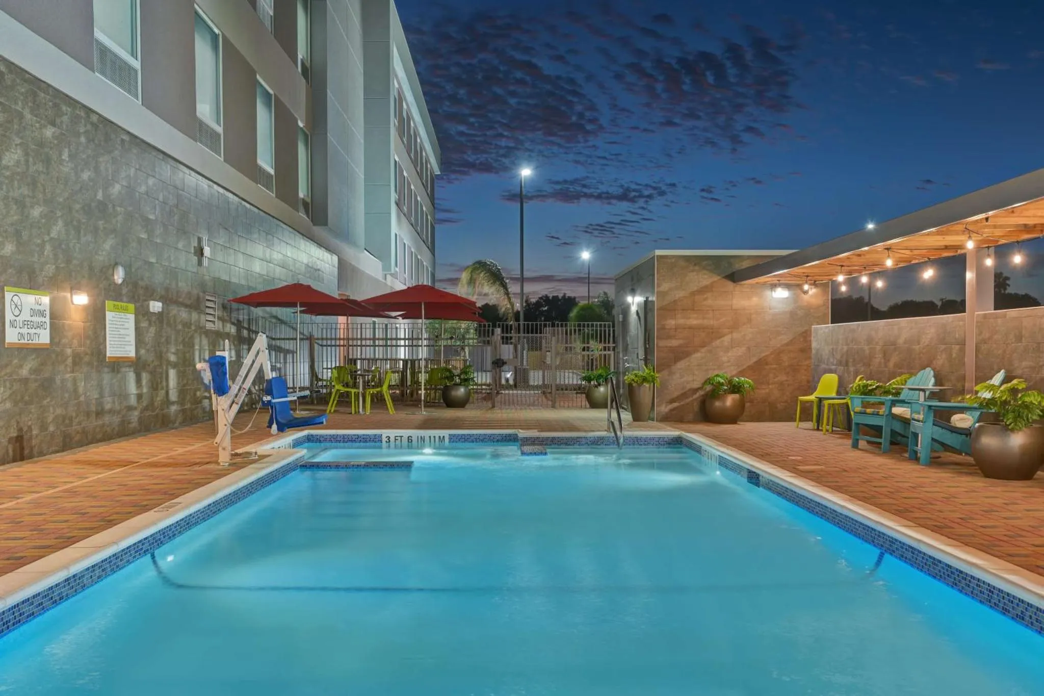 Une piscine rectangulaire avec des transats et des parasols, devant un bâtiment éclairé.