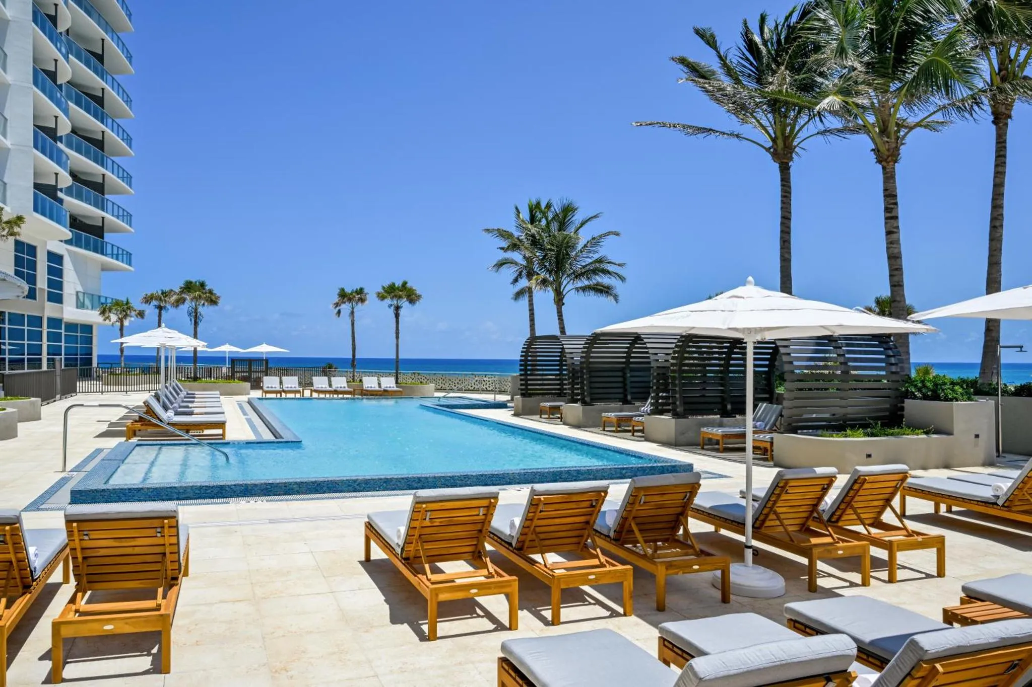 Transats et parasols bordent une piscine rectangulaire d'un hôtel en bord de mer.