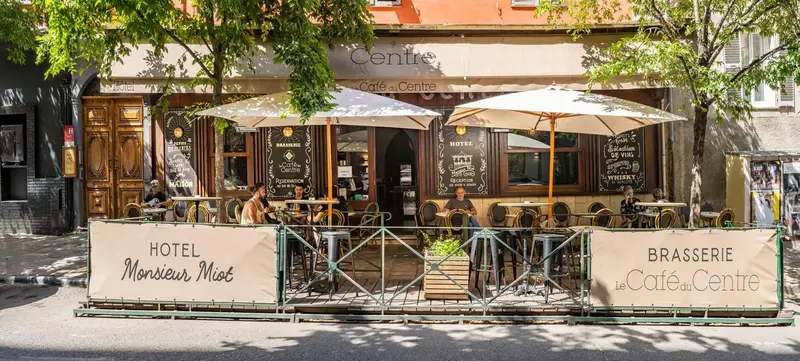 Terrasse de café avec tables et parasols devant la façade d'un hôtel et d'une brasserie.