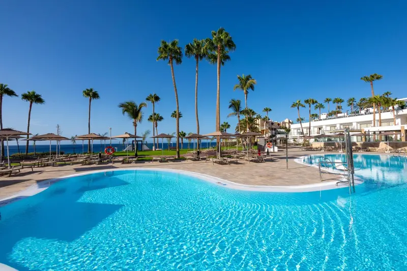 Piscine turquoise bordée d'une terrasse ensoleillée avec des palmiers et des parasols.
