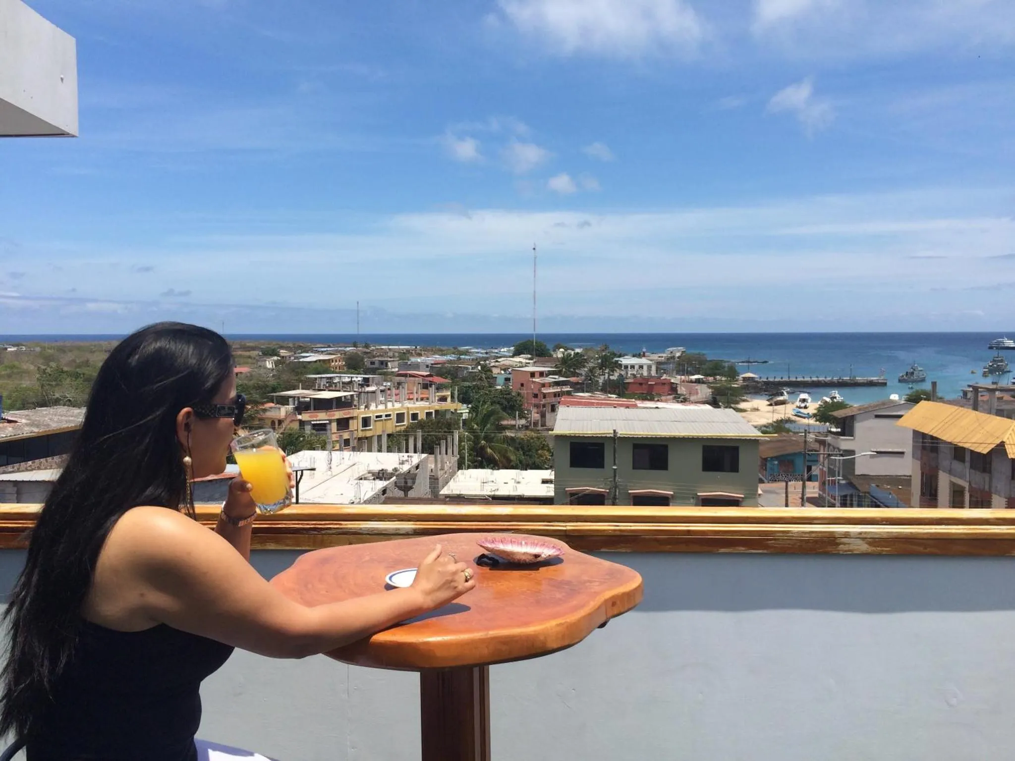 Une femme déguste une boisson sur une terrasse ensoleillée avec vue sur la ville et la mer.