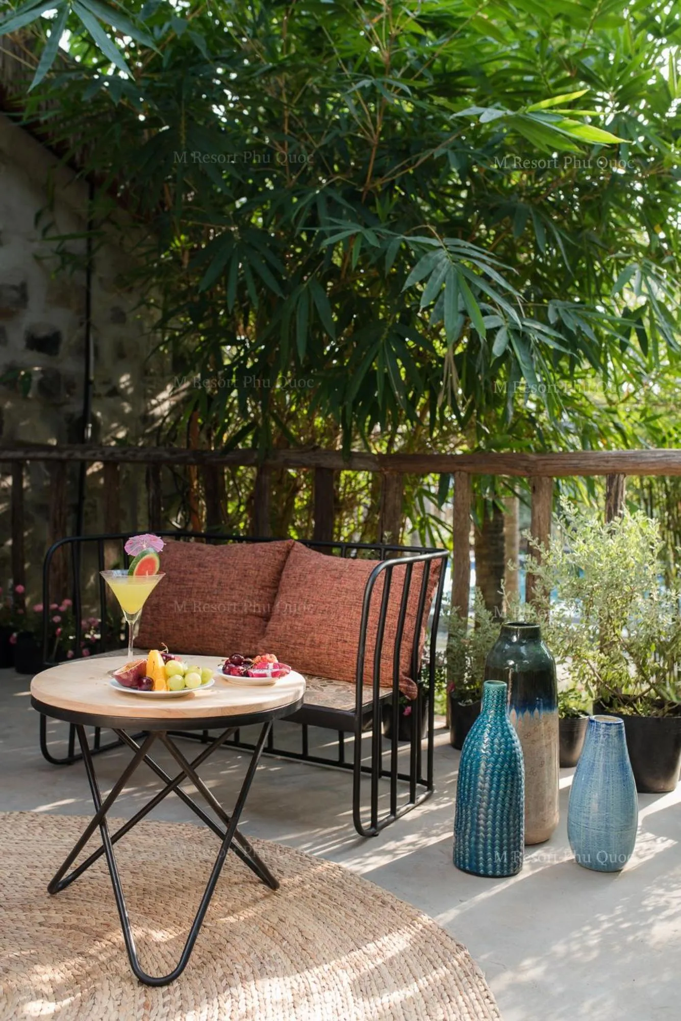 Une table basse avec des boissons et des fruits est placée devant un canapé sur une terrasse ombragée.