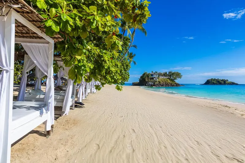 Transats blancs sous des pergolas sur une plage de sable fin.