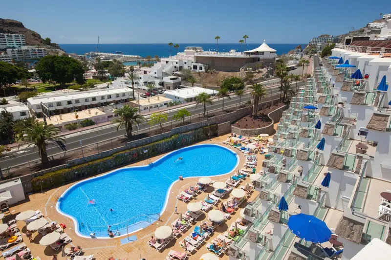 Vue aérienne d'une piscine d'hôtel avec des transats et des parasols, surplombant la mer et un port de plaisance.