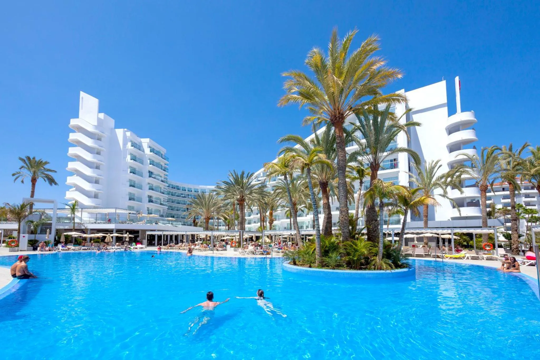 Vue d'une piscine d'hôtel bordée de transats et de palmiers sous un ciel bleu.