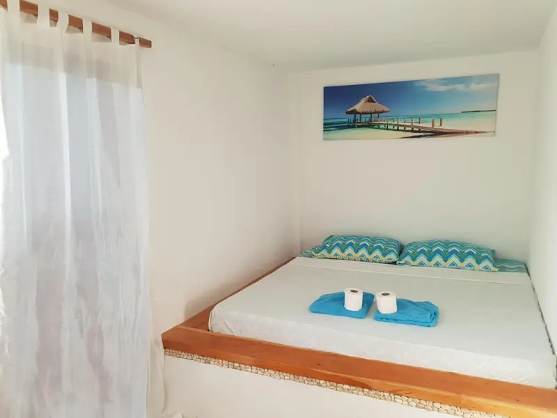 Une chambre lumineuse avec un lit décoré de coussins bleus et une grande toile représentant un paysage tropical.
