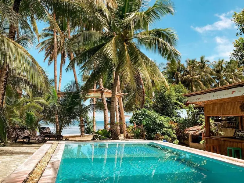 Une piscine turquoise bordée de chaises longues, avec des palmiers et des bâtiments en arrière-plan.