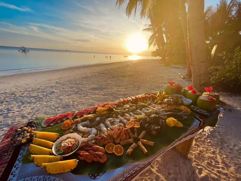 Une table basse garnie de fruits de mer et de fruits exotiques est dressée sur une plage de sable fin au coucher du soleil.