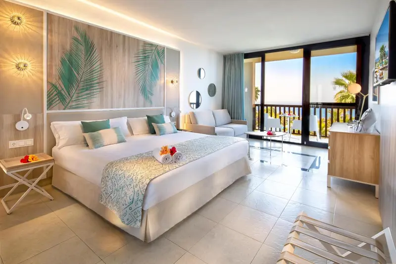 Chambre lumineuse avec un grand lit et un canapé face à une baie vitrée.