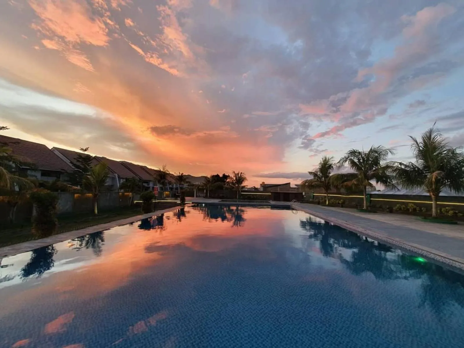Une piscine reflète un ciel orangé au coucher du soleil devant des bâtiments.