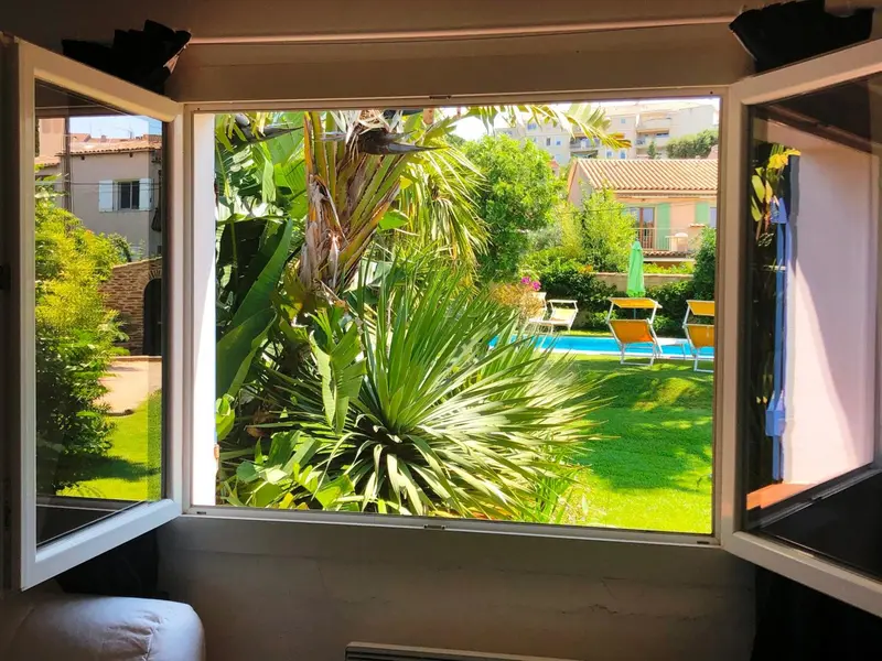 Vue sur un jardin verdoyant avec une piscine depuis une fenêtre ouverte.