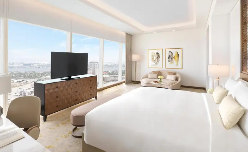 Chambre lumineuse avec un grand lit et un coin salon près d'une fenêtre panoramique.