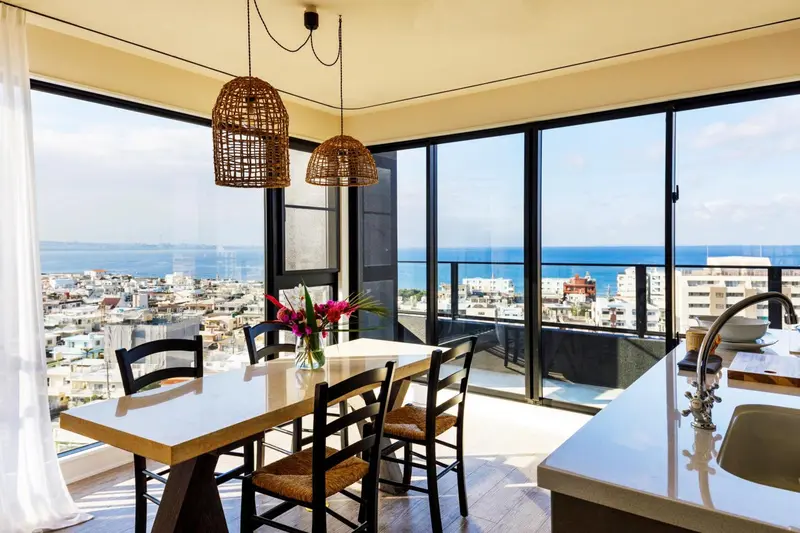Une table à manger en bois clair est mise en valeur par des luminaires suspendus dans une pièce lumineuse avec vue sur la mer.
