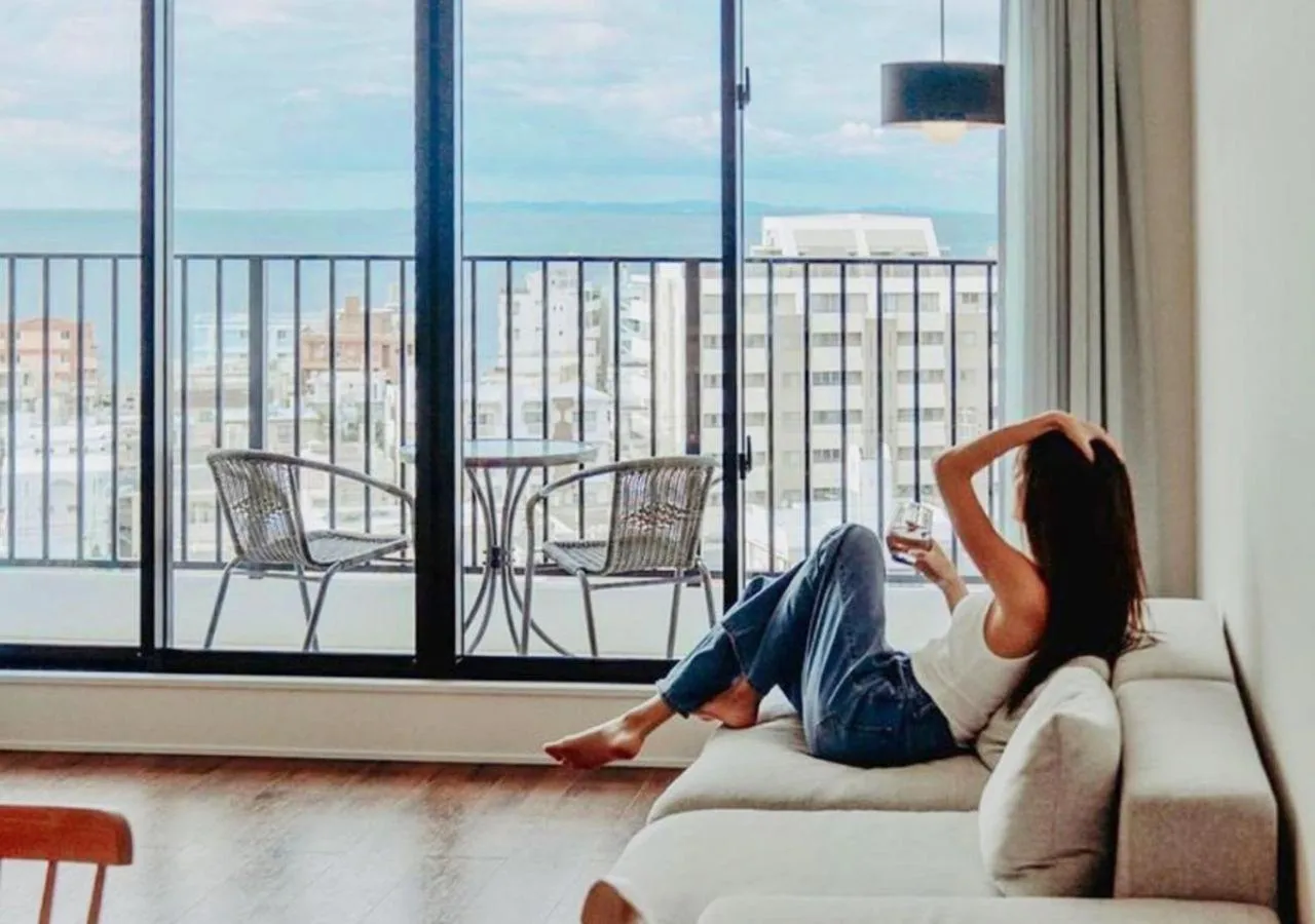 Une femme se détend sur un canapé avec une boisson, face à une vue sur la mer depuis un balcon.