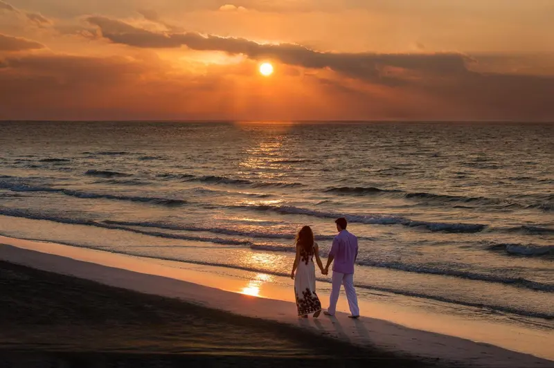 Un couple se tient par la main sur une plage au coucher du soleil.