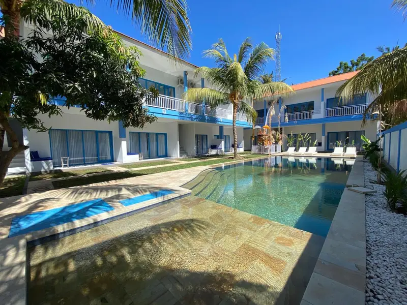Une piscine rectangulaire bordée d'une terrasse ensoleillée devant un bâtiment blanc à deux étages.