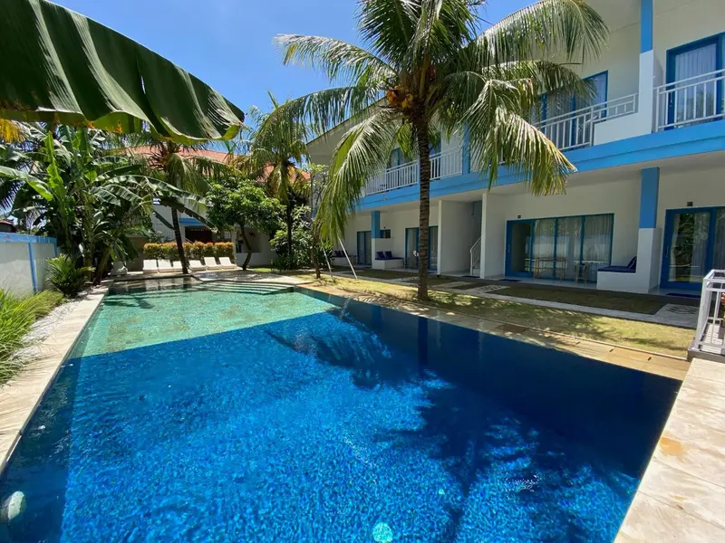 Une piscine rectangulaire turquoise est bordée d'une terrasse avec des chaises longues et des palmiers.