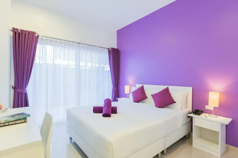 Une chambre lumineuse avec un grand lit recouvert d'une couette blanche et de coussins violets.