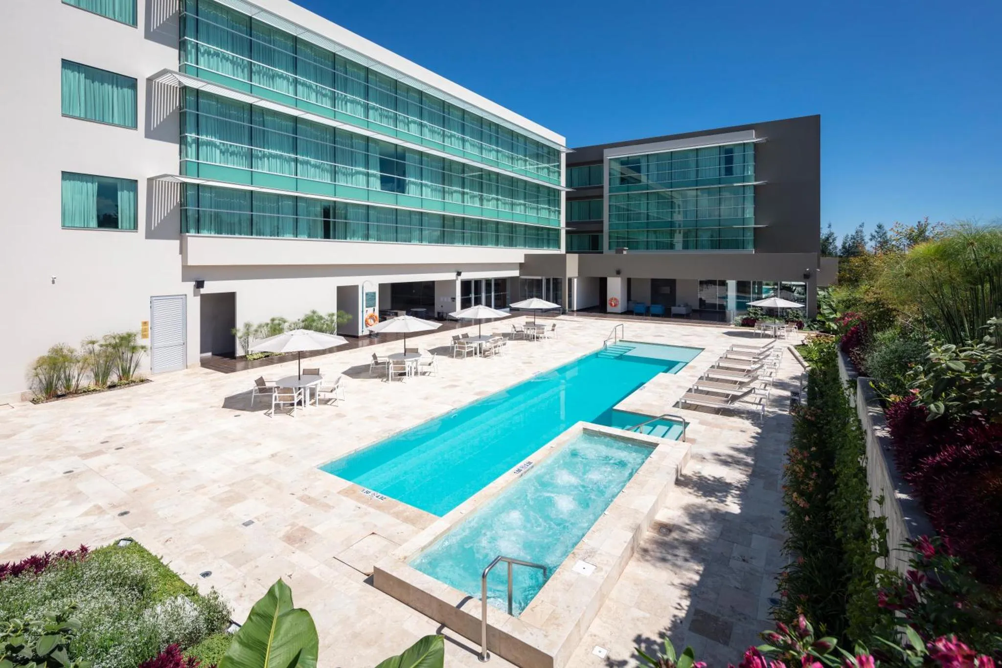 Piscine rectangulaire turquoise bordée de transats et de parasols devant un bâtiment moderne.