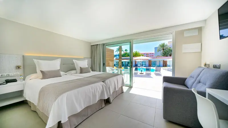 Une chambre lumineuse avec un grand lit et un coin salon donnant sur une piscine.