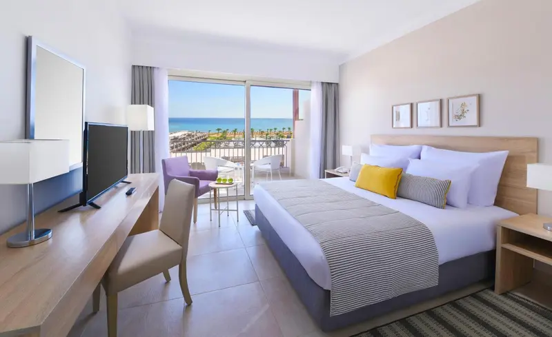 Chambre lumineuse avec un lit double et une vue sur la mer depuis un balcon.