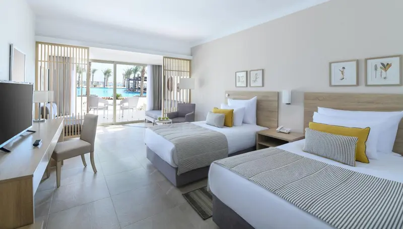 Chambre lumineuse avec deux lits et vue sur une piscine.