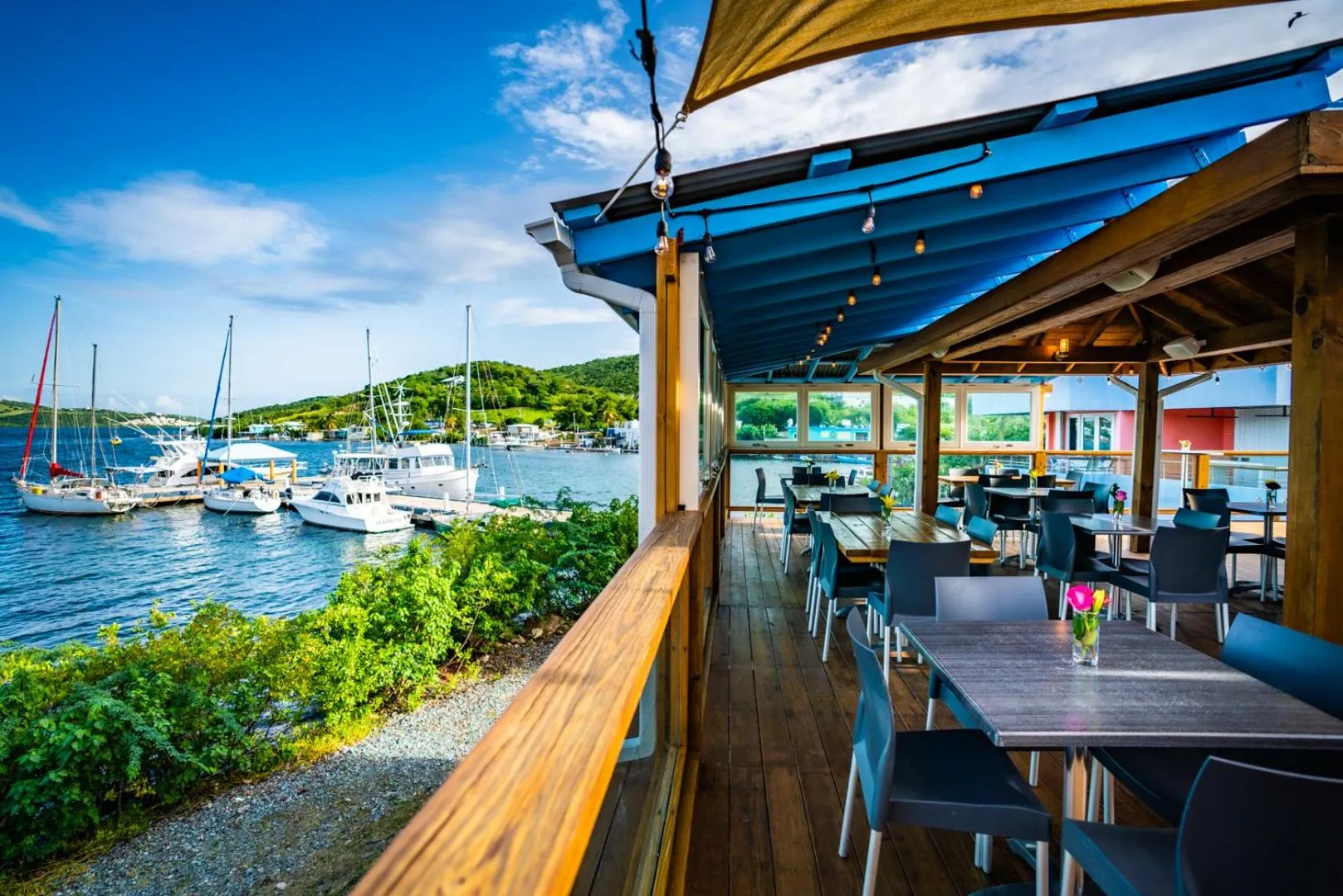 Terrasse ombragée avec tables et chaises donnant sur un port de plaisance.