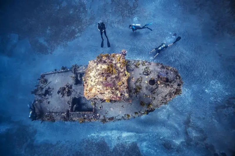 Vue aérienne d'une épave recouverte de corail, explorée par des plongeurs.