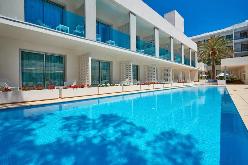 Piscine turquoise bordée de fleurs et de chaises longues devant un bâtiment blanc.