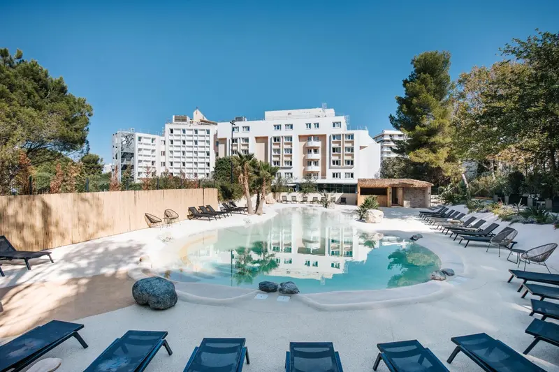 Une piscine bordée de transats et de végétation devant un bâtiment blanc.
